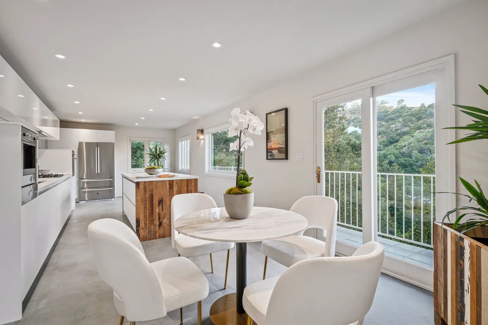 Kitchen and breakfast area with white chairs, opening to a balcony with tree views