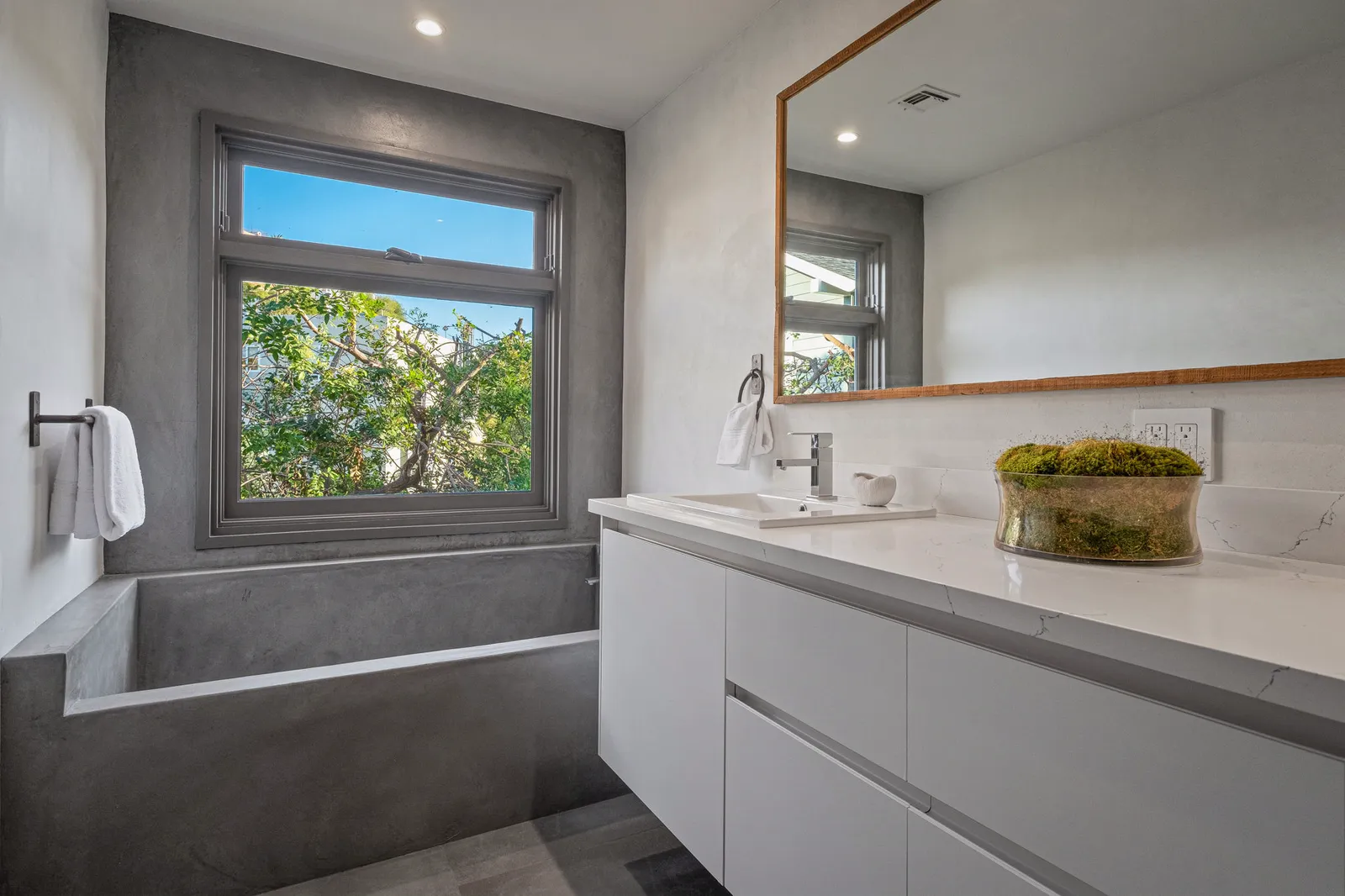 Bathroom with a gray concrete-look tub surround, white vanity, and a window to the trees