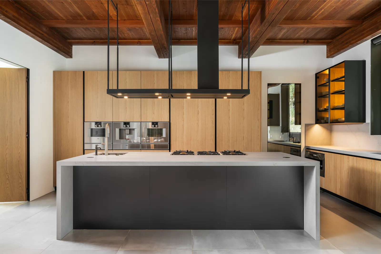 Kitchen with a dark island and waterfall stone top, wood cabinetry, and a black ceiling hood