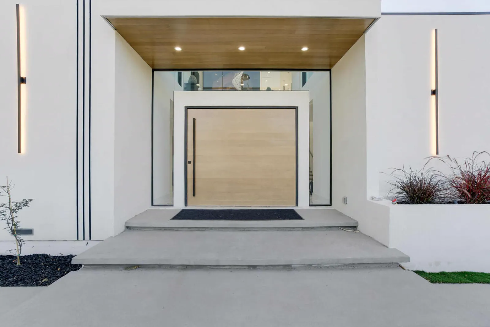 Entry with a tall wood pivot door under a lit wood soffit