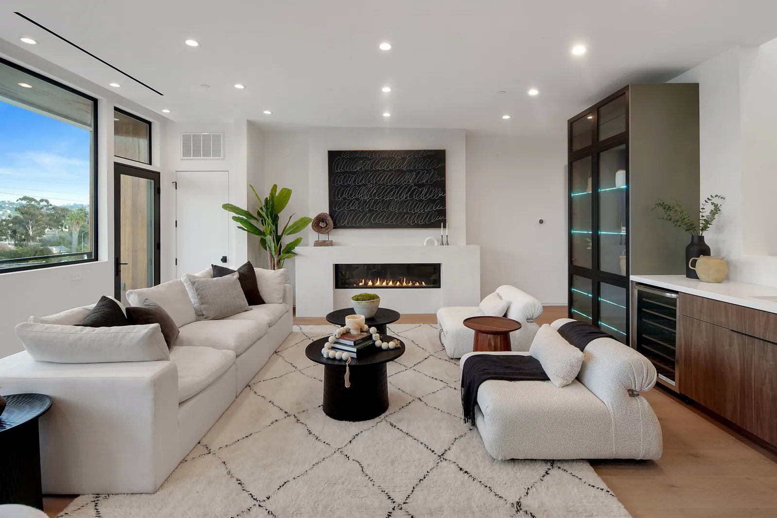 Living room with white sectional sofas, a linear fireplace, and a glass-front bar cabinet beside a large window