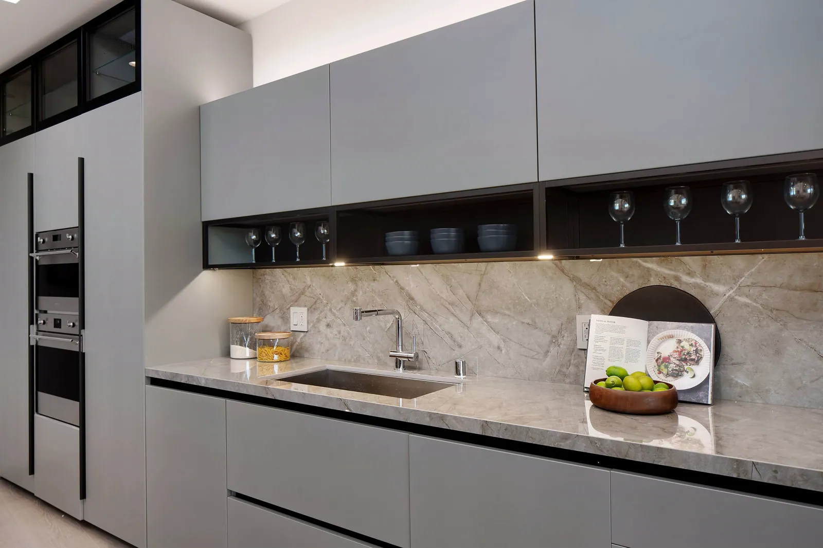 Kitchen counter with green-gray upper cabinets, built-in ovens, and a stone backsplash