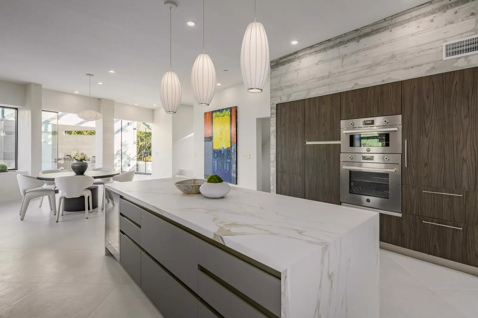 Long marble kitchen island beside dark wood tall cabinets and built-in ovens