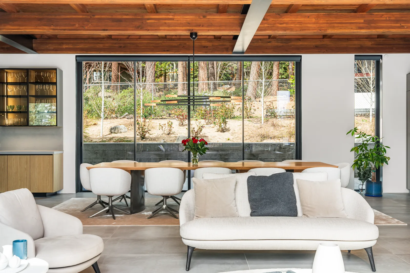 Living area with white sofas, a long dining table, and a wall of glass to the forest